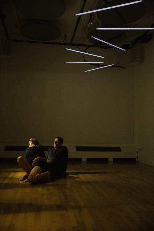 Two dancers sit in a dark room. 5 strips of lights cast a small pool light on them